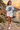 Female model wearing the light-blue skull-skate tee beside a Mediterranean cove