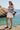 Full-length female model wearing the pastel striped all-over-print sweatshirt with tailored white shorts at a coastal cove