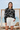 Full-length female model wearing the black botanical all-over-print sweatshirt with tailored white shorts beside the ocean