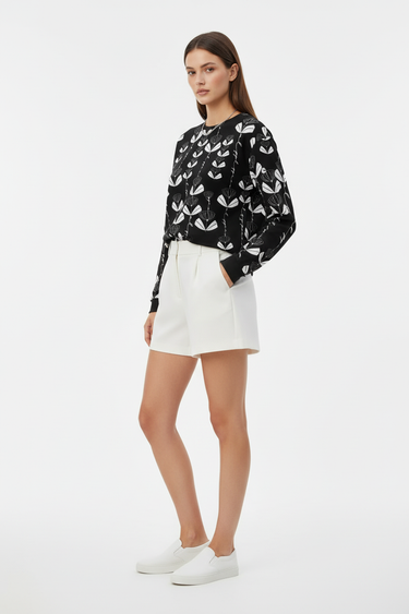 Full-length female model wearing the black botanical all-over-print sweatshirt with white tailored shorts on a white background, side view