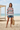 Full-length female model wearing the pastel striped all-over-print sweatshirt with vintage denim shorts beside the ocean