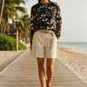 Full-length female model wearing the black botanical all-over-print sweatshirt with tailored cream Bermuda shorts on a beach boardwalk