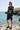Full-length female model wearing the black botanical all-over-print sweatshirt with tailored navy shorts at a coastal cove