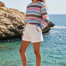 Full-length female model wearing the pastel striped all-over-print sweatshirt with tailored white shorts at a coastal cove