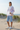 Full-length female model wearing the pastel all-over-print recycled hoodie with tailored pale blue shorts among coastal dunes