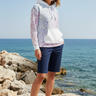 Full-length female model wearing the pastel all-over-print recycled hoodie with tailored navy shorts at a coastal cove
