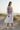 Full-length female model wearing the pastel striped all-over-print sweatshirt with a white midi skirt among coastal dunes