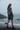 Full-length female model wearing the black botanical all-over-print sweatshirt with tailored charcoal shorts on a Pacific shoreline