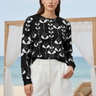 Full-length female model wearing the black botanical all-over-print sweatshirt with tailored white shorts beside the ocean